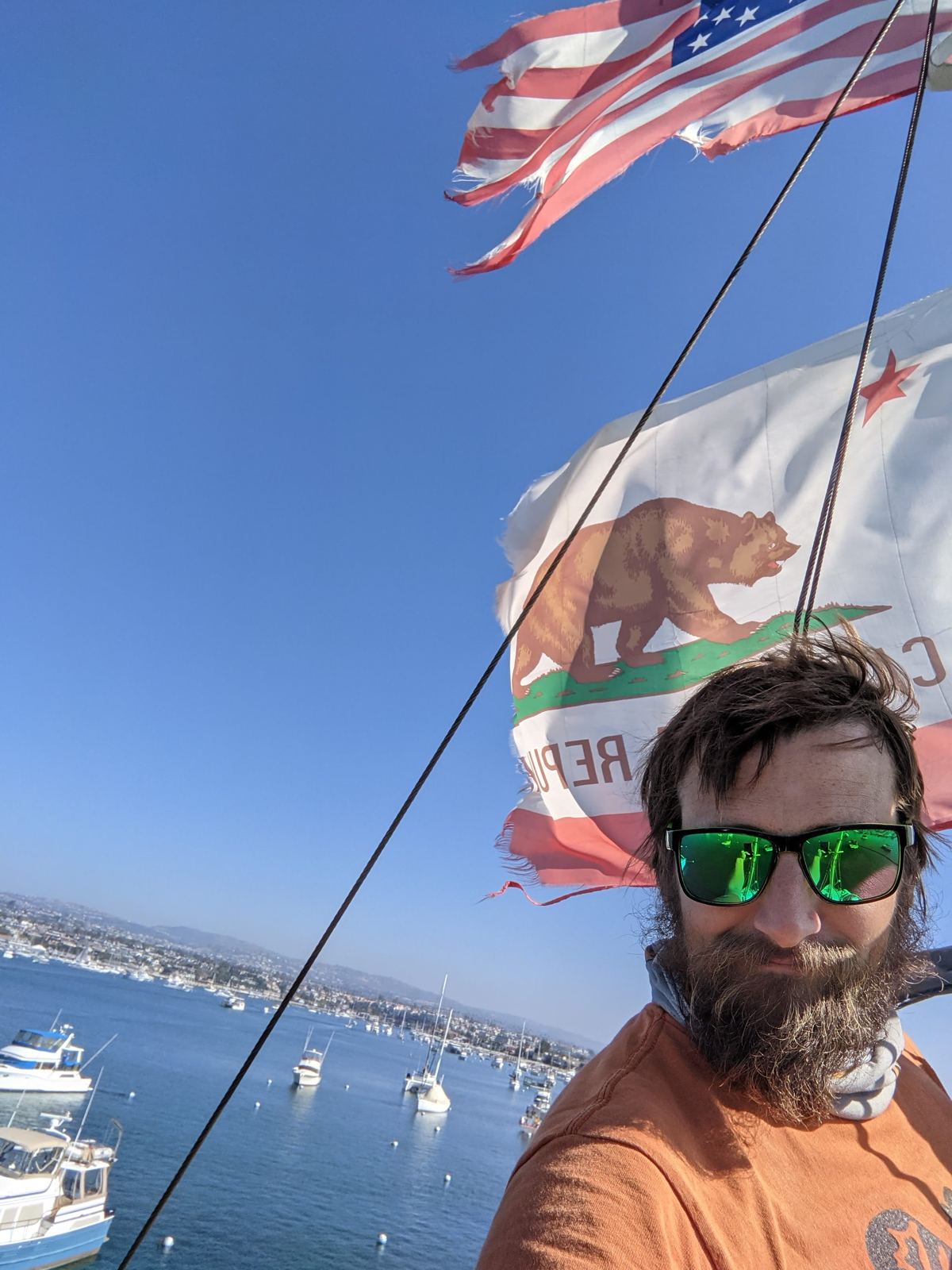 Brian aloft in Windance's rigging, flags flying, with Newport Harbor below