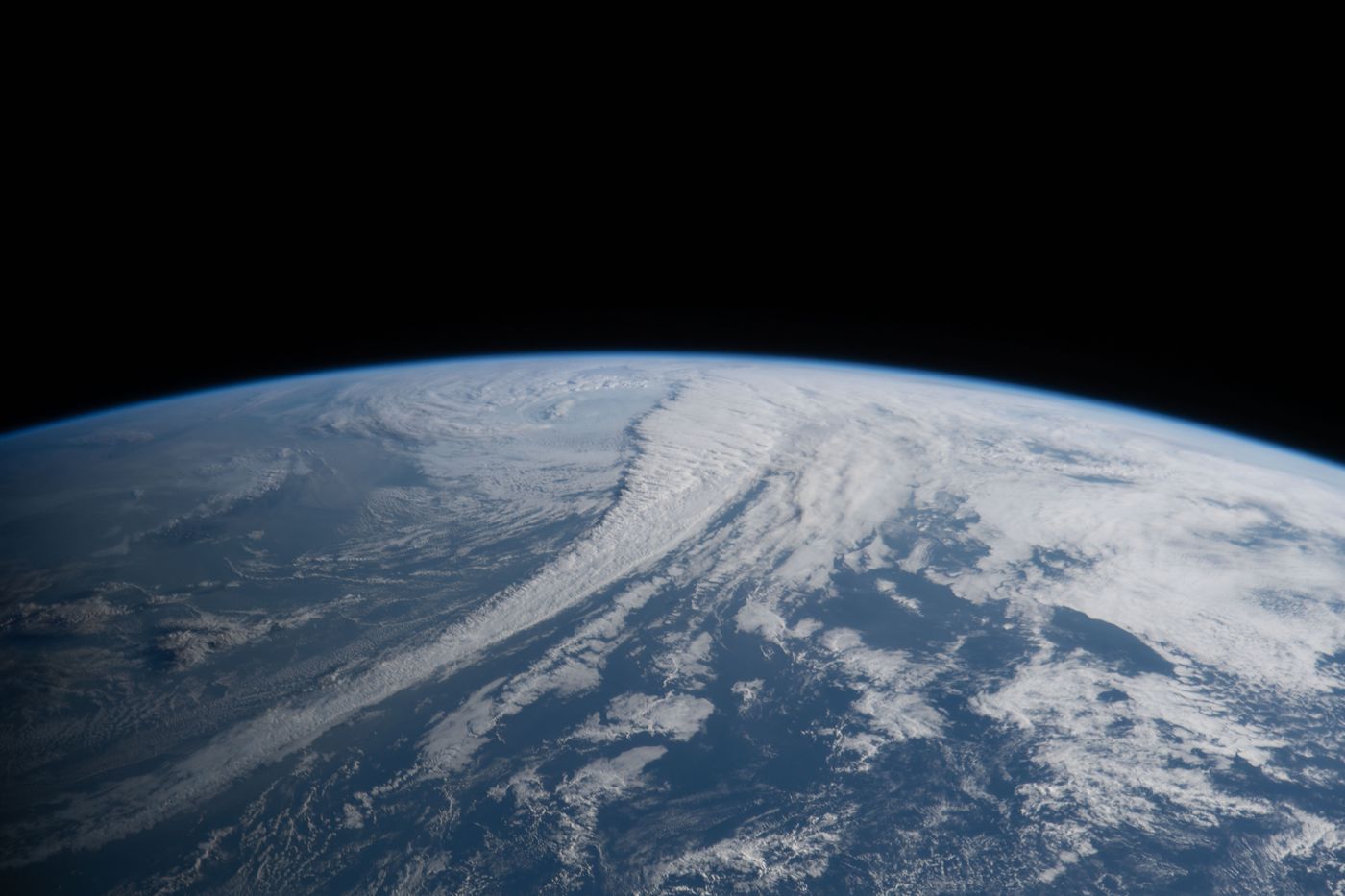 The curved limb of Earth with long bands of white cloud over blue ocean beneath the black of space